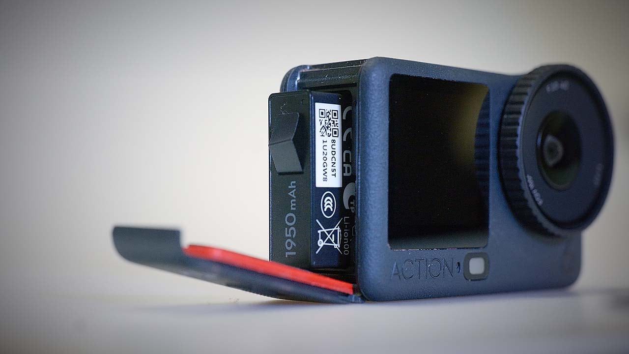 The image features the DJI Action 6, partially opened to reveal its battery compartment. The camera has a sleek design with a prominent lens on the front and a textured grip. The battery is labeled with specifications, including its capacity of 1950mAh. The background is softly blurred, emphasizing the camera.