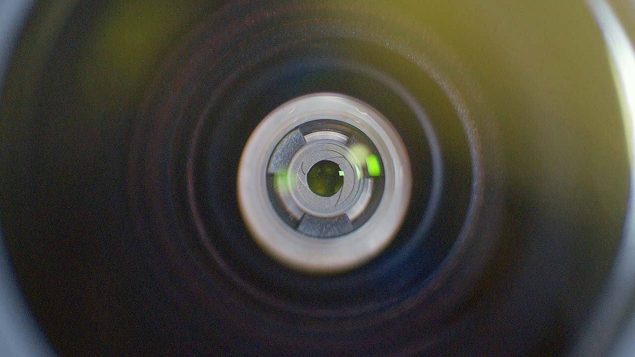 The image features a close-up view of the DJI Action 6 lens, showcasing the intricate details of the lens elements and the aperture. The circular shape and layered design highlight the optical components, with reflections of light visible, indicating the lens's functionality in capturing images. The background appears blurred, emphasizing the lens itself.