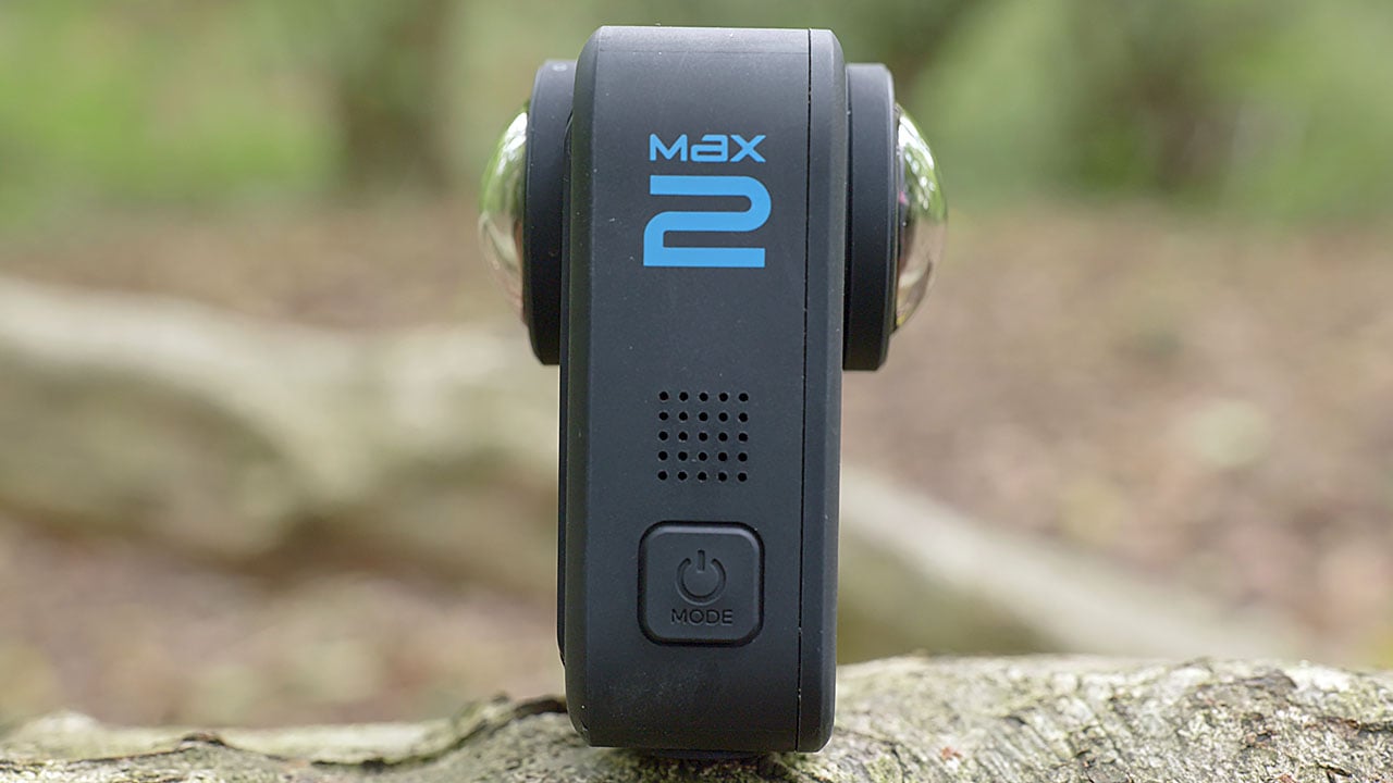 The image shows a small, compact device with a black casing and the brand name 'Max' in white letters. It has a '+' button, a speaker, and a green button labeled 'MODE'. The device is placed on a wooden surface with a blurred natural background, suggesting an outdoor setting.