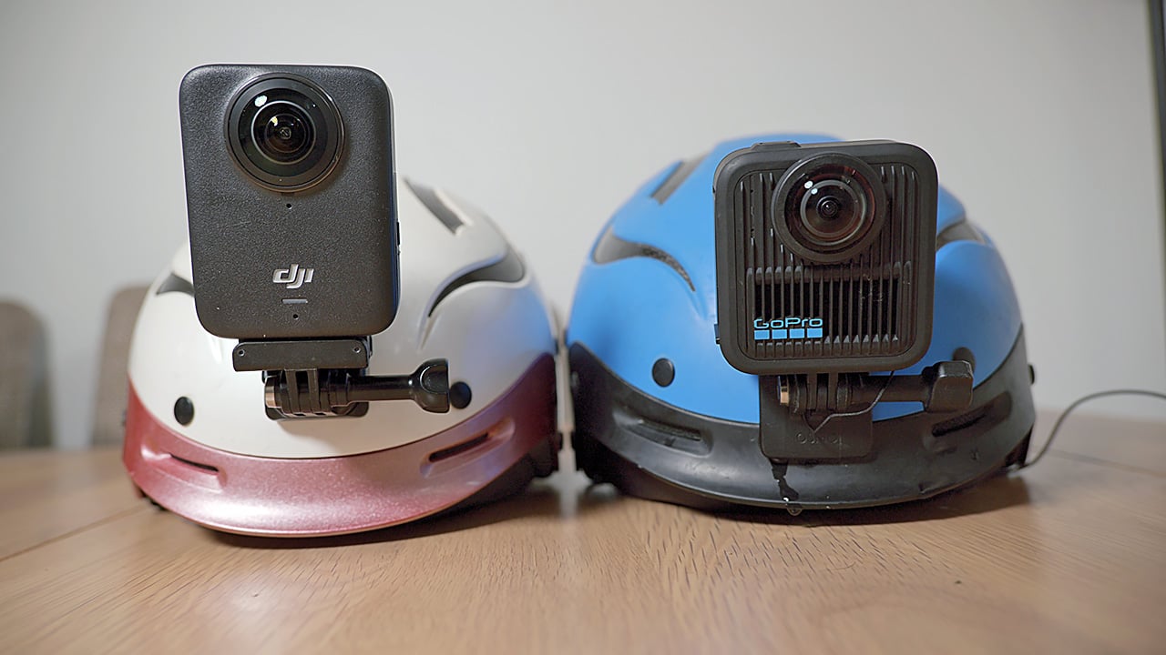 The image shows two kayaking helmets with camera mounts. On the left, there is a helmet with a DJI Osmo 360 mounted on its top, and on the right, there is a helmet with the GoPro Max 2 mounted on its top. The DJI camera is noticeably taller. Both helmets are placed on a wooden surface, and the one on the right has a blue body, while the one on the left has a white and red body.