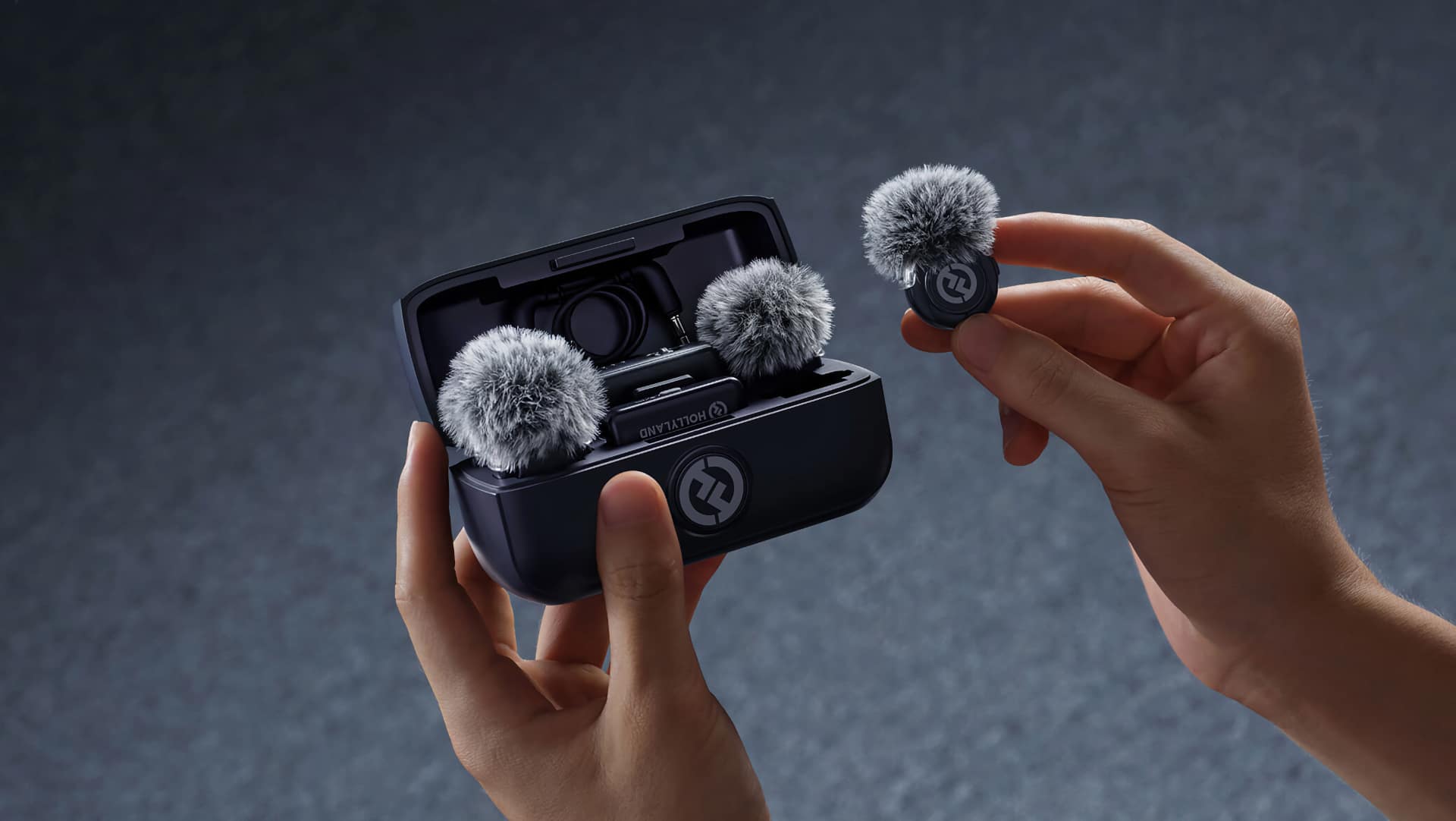 Hollyland LARK M3 charging case open in one hand, showing two furry windshields and a transmitter, with a third transmitter held in the other hand