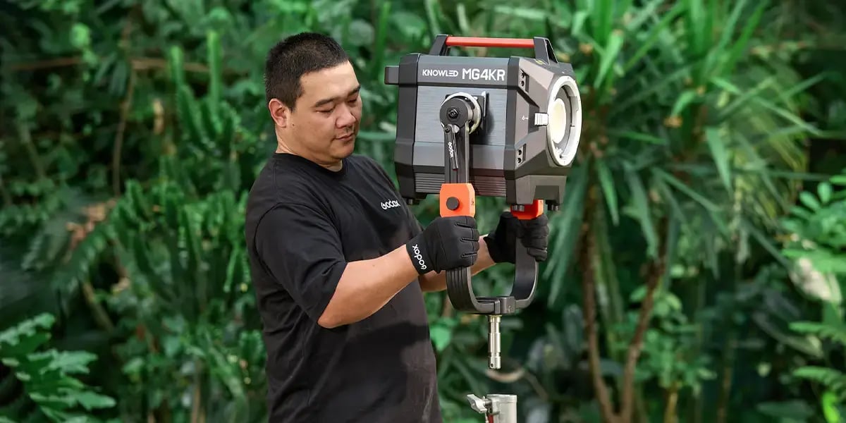 The image features a man operating a large, professional-grade lighting equipment, specifically the KNOWLED MG4KR model. He is wearing black gloves and a casual black shirt, with a backdrop of lush green foliage, suggesting an outdoor or studio setting that emphasizes greenery. The man appears focused on his task, indicating a professional or technical environment related to photography or film production.