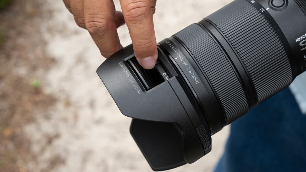 A hand inserts a square lens filter into the slot of a NIKKOR Z 24-70mm f/2.8 S II  lens hood attached to a black camera lens, outdoors on a blurred natural background.