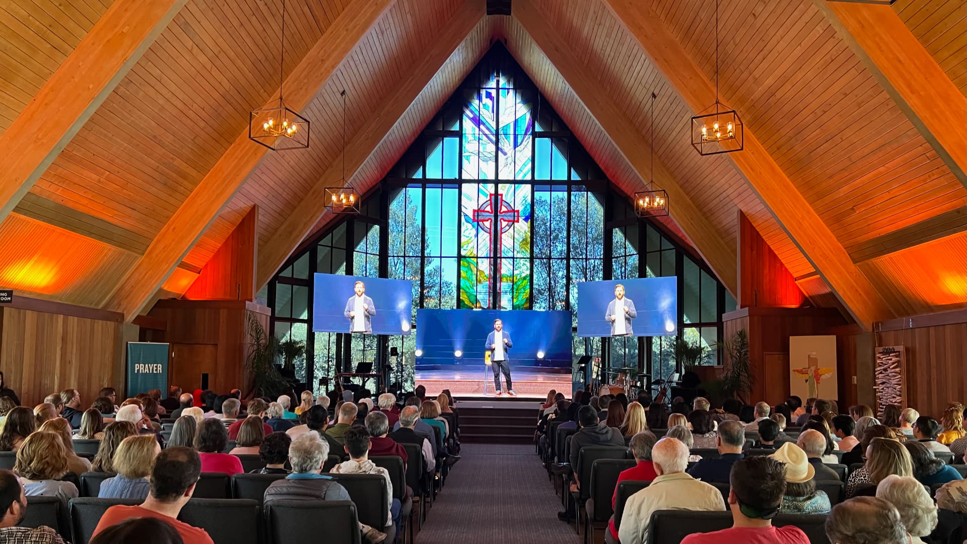 The image depicts a church service taking place inside a building with a large stained glass window at the front. The room is filled with an audience facing a stage where two individuals are presenting, with large screens displaying their image.