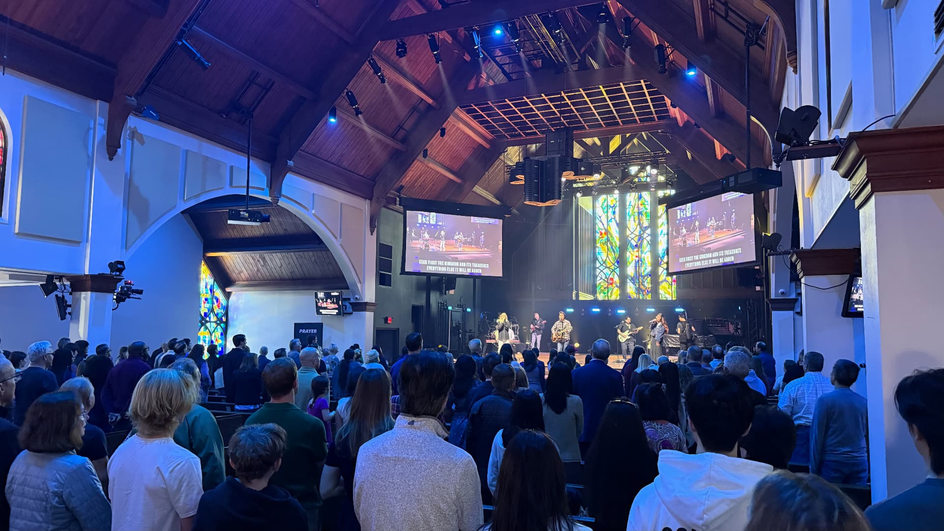 The image shows a church service with a band performing on stage in front of a large audience. The audience is seated facing the stage, and there is a stained glass window in the background. The room has wooden walls and a high ceiling, and there are lights and a large screen visible on stage.