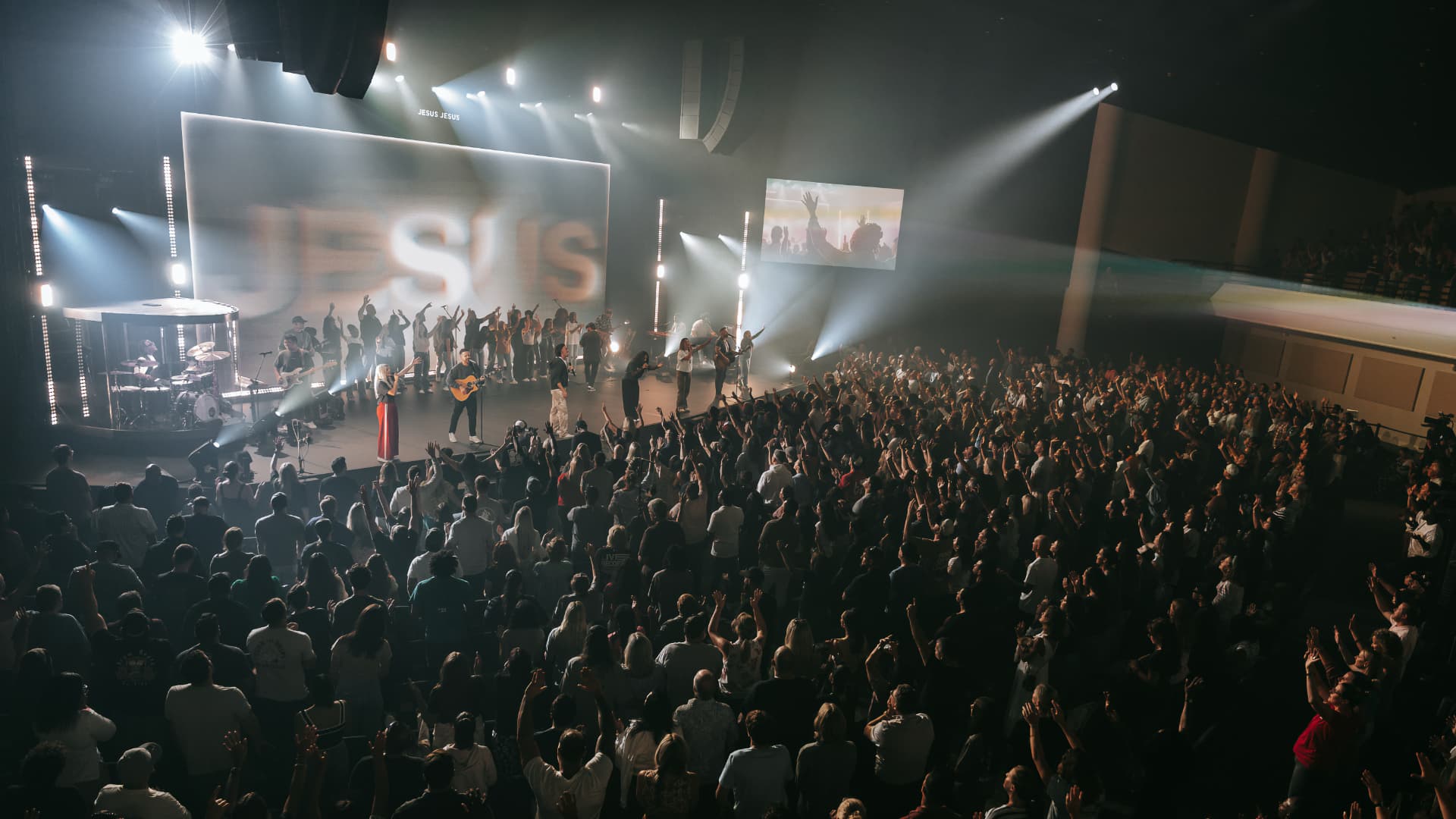 The image captures a vibrant scene from a concert or event. The stage, bathed in the glow of bright lights, is adorned with a large projection displaying the word "JESUS". The audience, a sea of people, fills the space, their attention riveted on the stage. The perspective is from the side, looking towards the stage, creating a sense of anticipation and excitement. The image is in black and white, adding a dramatic effect to the scene.