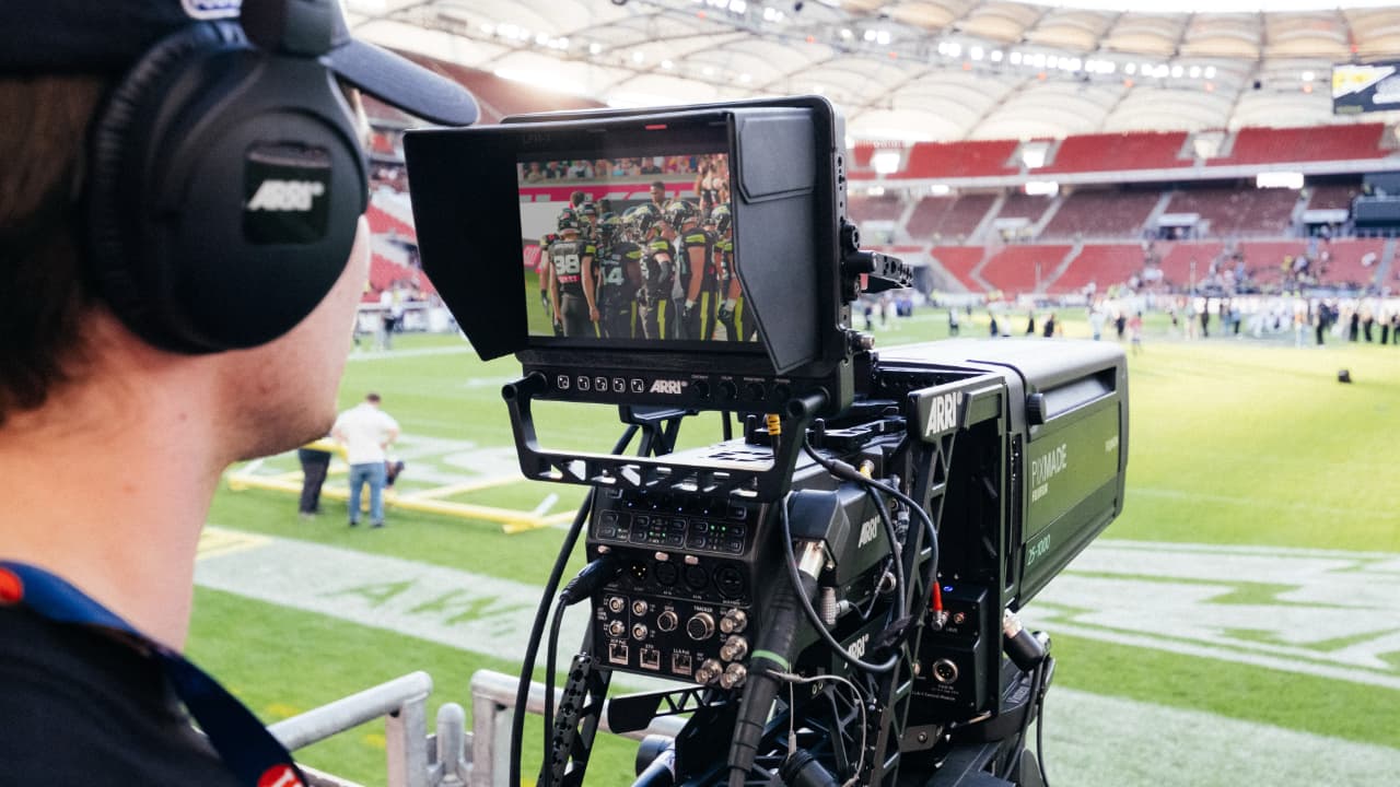 The image shows a professional video camera operator using a camera positioned on a tripod, equipped with headphones, focused on capturing an event in a stadium. The camera is recording a scene of a sports event, specifically what appears to be a soccer game, with players on the field and spectators in the stands.