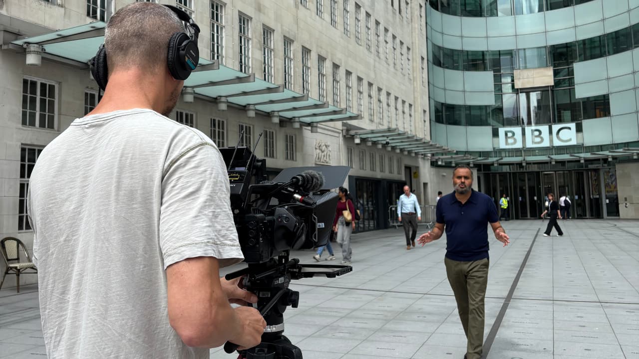 The image shows a professional filming setup with a camera operator wearing headphones and holding a camera on a tripod, filming a scene in front of a building with the BBC logo. A man in a blue shirt is walking behind the camera, and there are other individuals in the background.