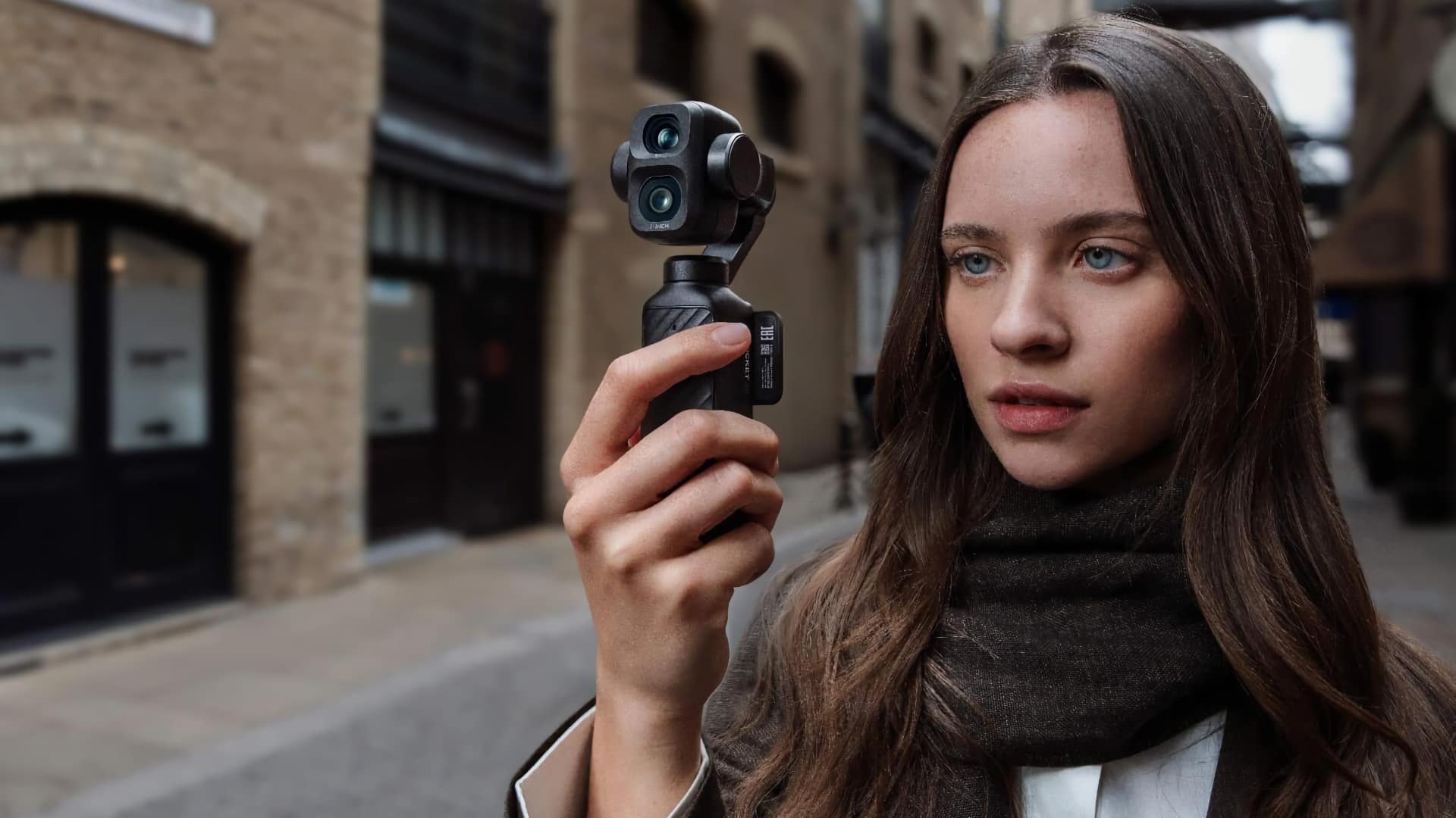 DJI Osmo Pocket 4P held in one hand against a blurred urban street backdrop, dual-lens gimbal head visible
