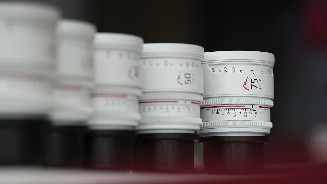 The image shows a close-up of a series of camera lenses or lens filters aligned horizontally. Each lens or filter has a white label around the edge with red text indicating the focal length and some have a red and black logo with 'CINE' visible. The background is blurred, emphasizing the lenses in the foreground.