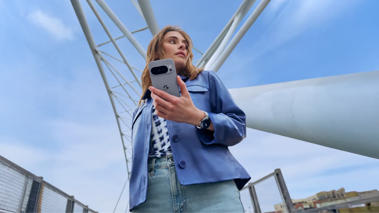 A woman in a blue jacket and jeans stands outdoors on a modern bridge, holding a smartphone and looking to the side under a blue sky with some clouds. The photo is taken from a low angle.