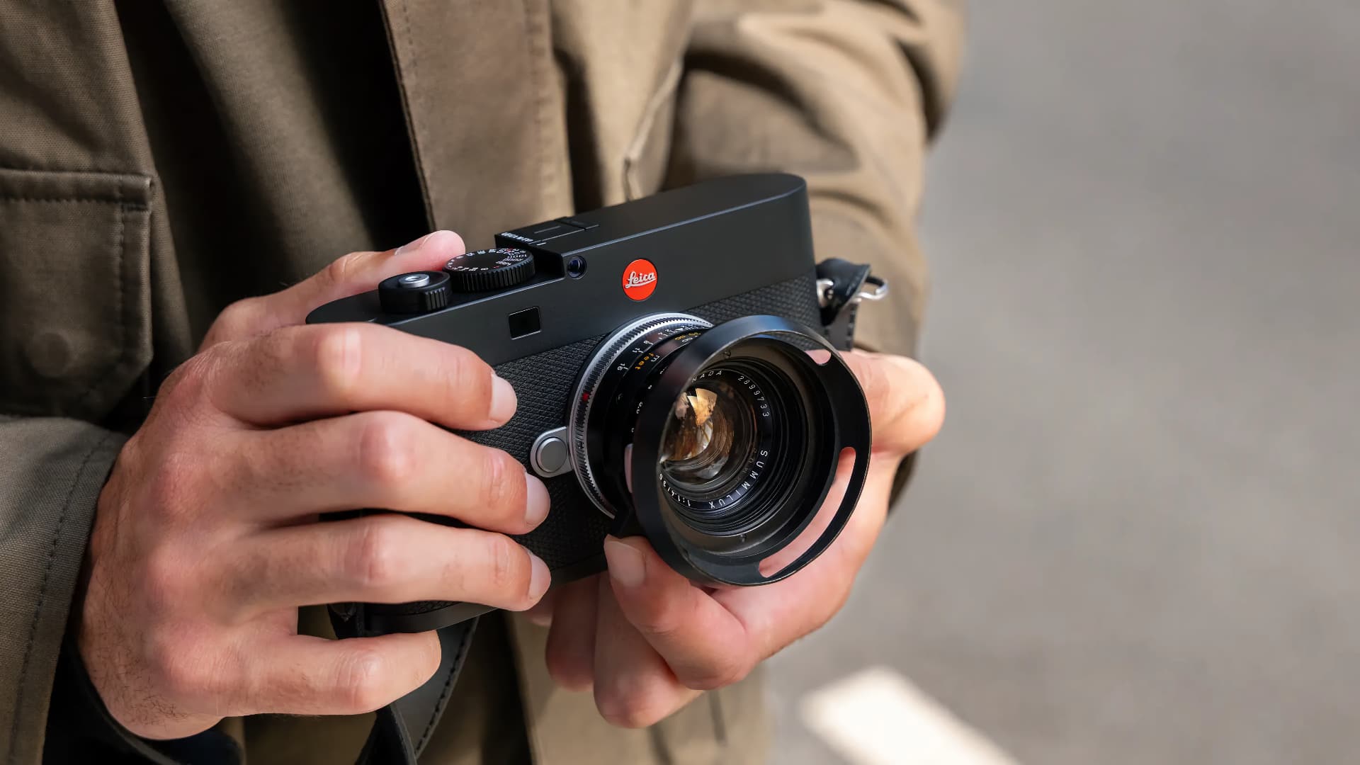 The image shows a person holding a Leica M EV1 camera with a prominent lens and a logo on the front. The person is wearing a brown jacket, and the background is blurred.