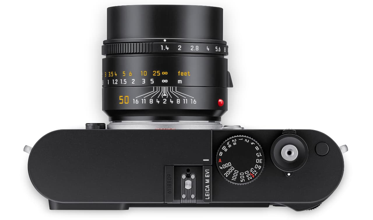 The image shows a close-up of a Leica M EV1 with an attached lens. The camera features a dial with numerical markings, aperture settings printed on the lens, and a viewfinder screen. The lens has a focal length of 50mm.