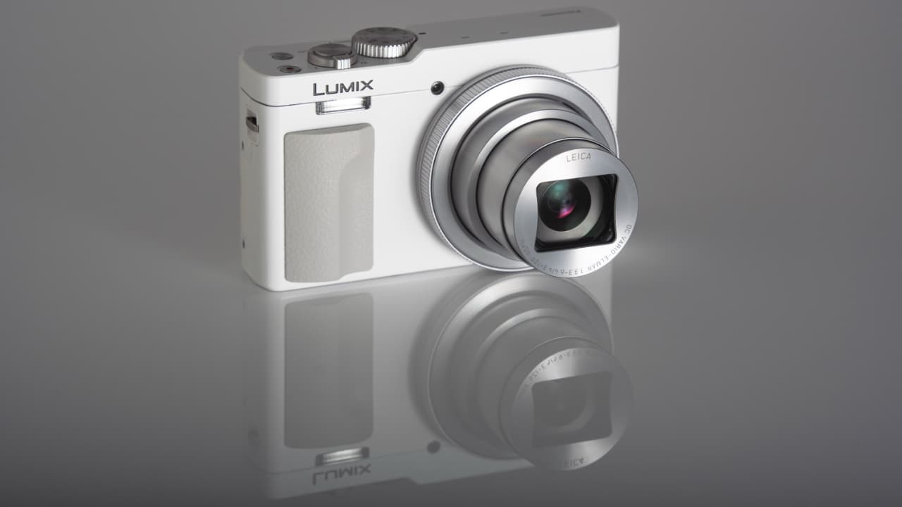 The image shows a Panasonic LUMIX TZ99 camera placed on a reflective surface. The camera is white in color with the brand name 'Panasonic' and model 'LUMIX' visible on its body.