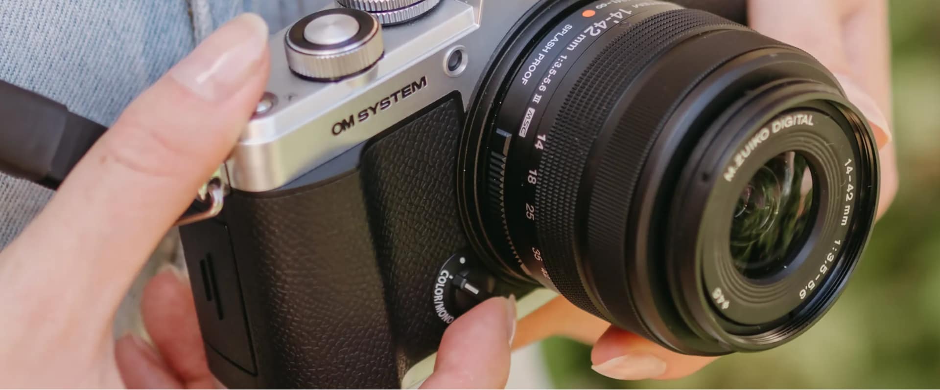 OM System PEN held in hand, showing the color/mono switch and 14-42mm splash-proof kit lens